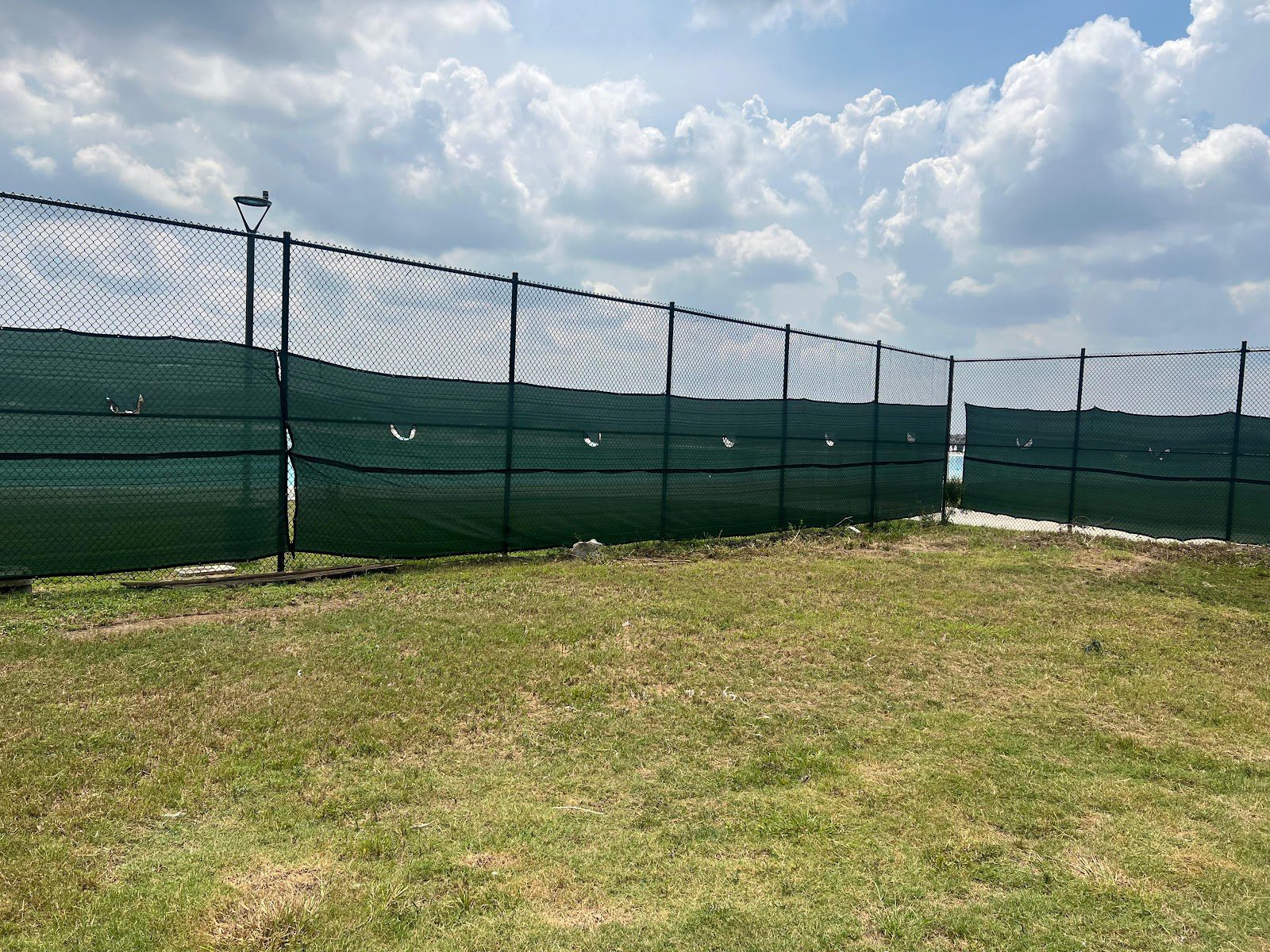 Corner section of black chain-link fence with green privacy windscreen and a light pole on a grassy commercial lot