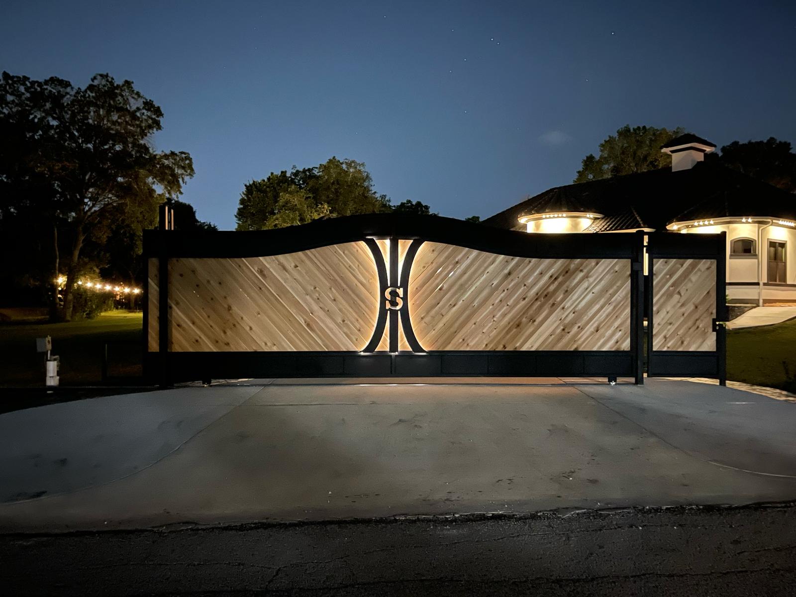 Custom cedar-and-black-steel driveway entrance gate with an illuminated monogram medallion at dusk
