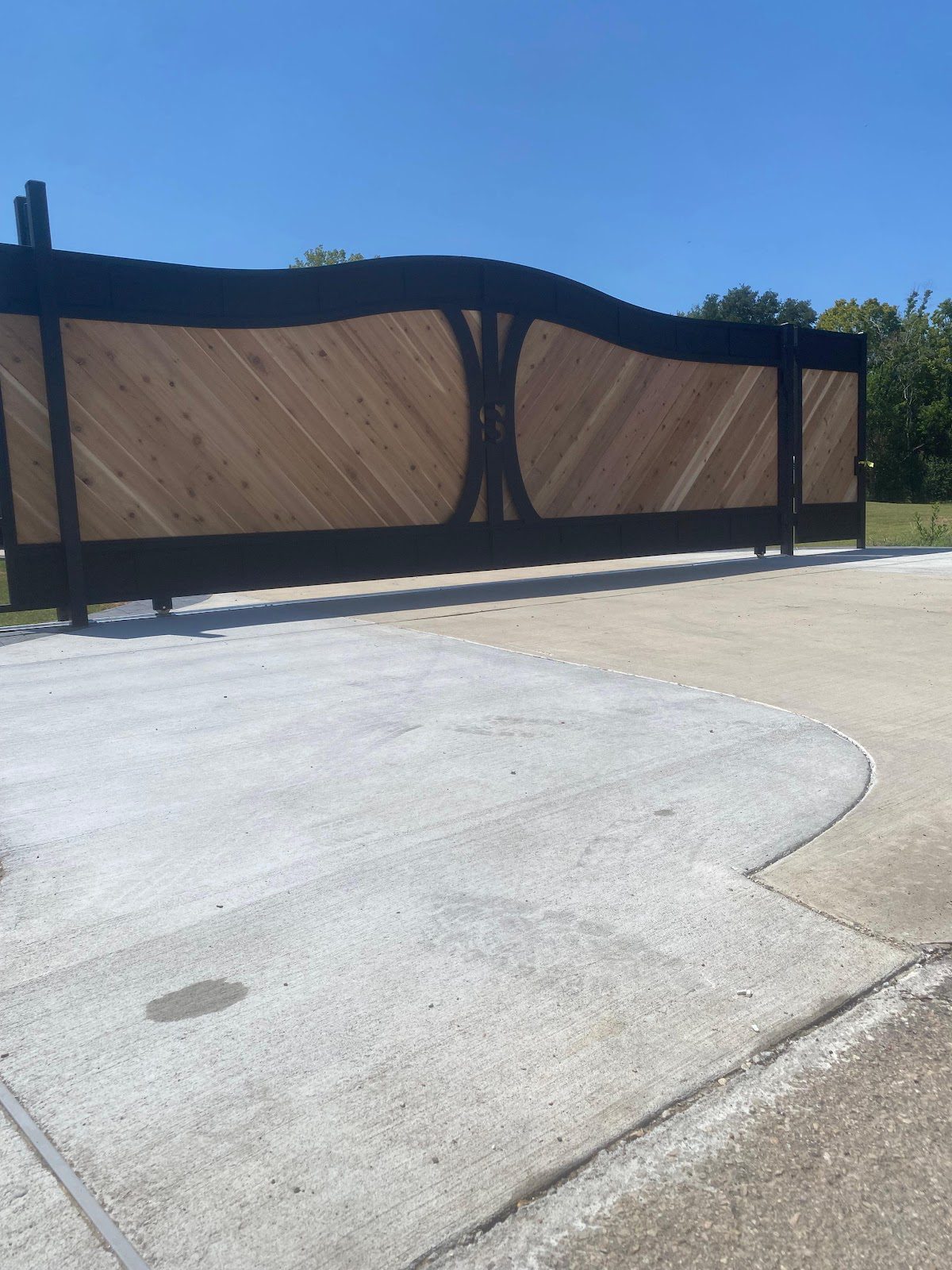 Wide view of a custom steel-and-cedar sliding driveway gate with a wavy top rail on a Houston-area property
