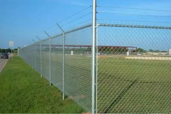 Galvanized chain-link perimeter fence topped with angled barbed wire screening an industrial site along a roadway