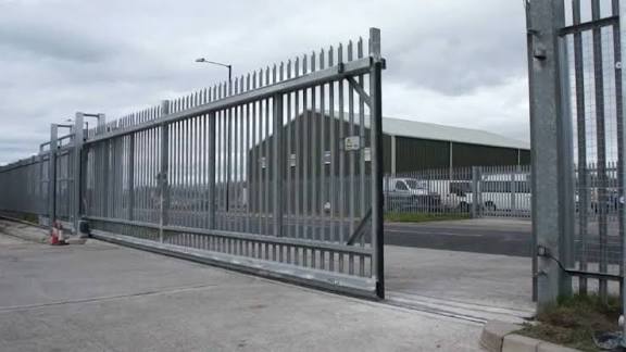 Galvanized steel palisade cantilever slide gate at the entrance to an industrial estate with a warehouse behind it
