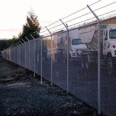 Galvanized chain-link fence with barbed wire top enclosing a commercial service yard with fleet trucks at dusk