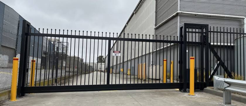 A bold black steel spear-top security slide gate flanked by yellow safety bollards, guarding the drive of a metal-clad commercial warehouse.