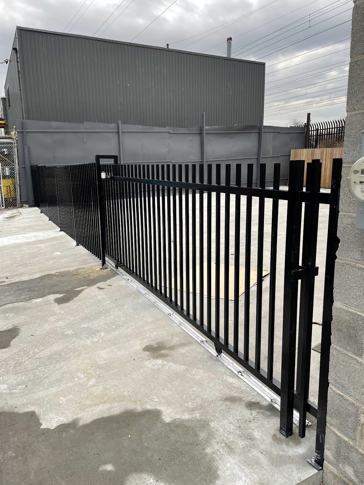 A heavy-duty black steel cantilever slide gate riding its track at a commercial industrial facility, framed by a metal building and privacy-screen wall.