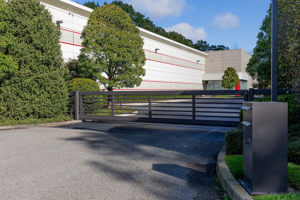 A keypad and call-box pedestal controls entry through a modern black horizontal-slat sliding gate at a landscaped commercial office campus.