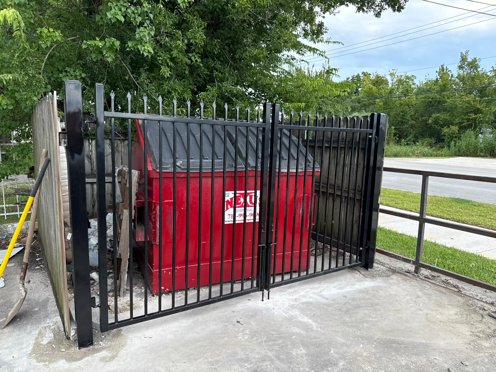A heavy black steel spear-top double swing gate closing off a commercial dumpster enclosure around the trash bin on a concrete pad.