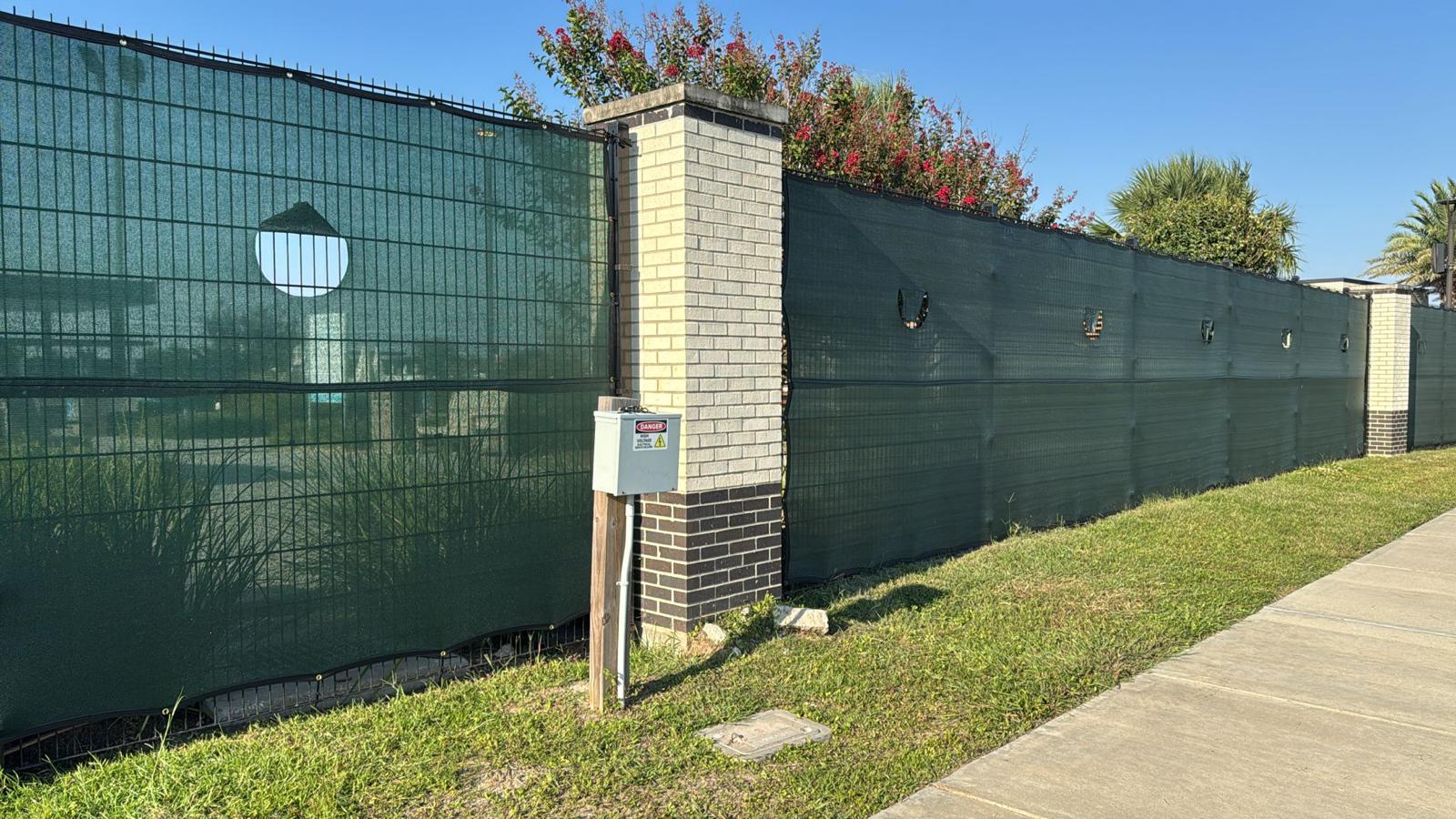 Black welded twin-wire mesh panels backed with green privacy screening enclose a commercial frontage, with a high-voltage utility box mounted in front. This rigid, tamper-resistant mesh is ideal for shielding utility service yards and equipment areas in greater Houston.