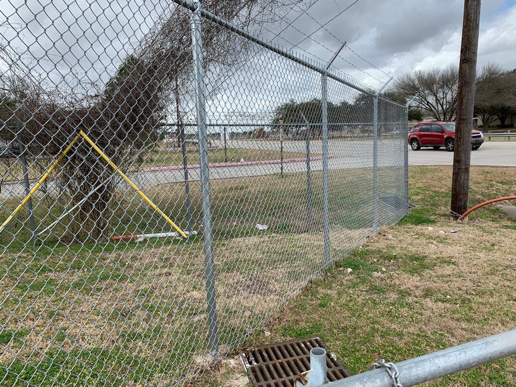 A galvanized commercial chain-link perimeter topped with a three-strand barbed-wire outrigger securing a roadway and parking area.