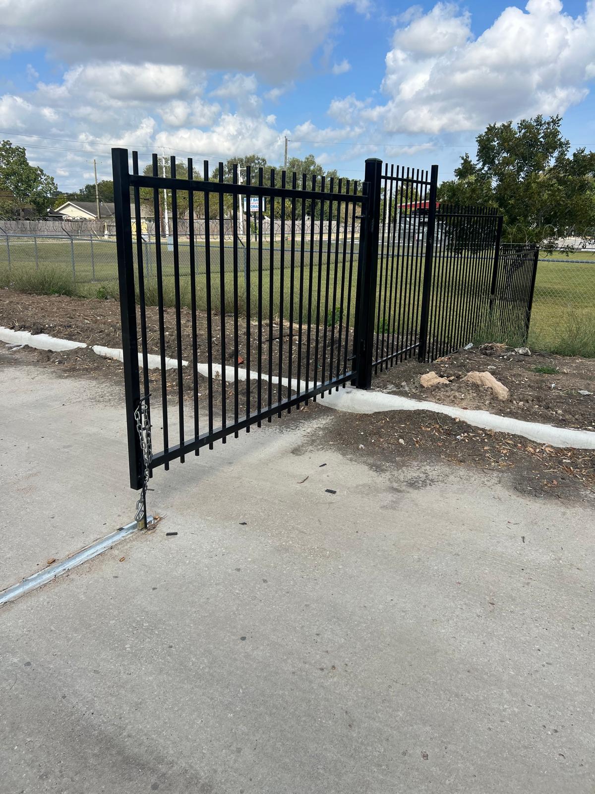 A black steel spear-top single swing gate stands open on its hinge post, tied seamlessly into a matching steel security fence across a commercial perimeter.