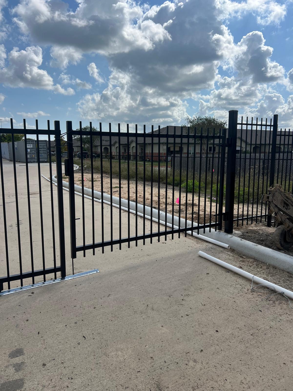 A black spear-top steel double swing gate stands open across a commercial drive, its two matched leaves swinging clear to admit vehicles.