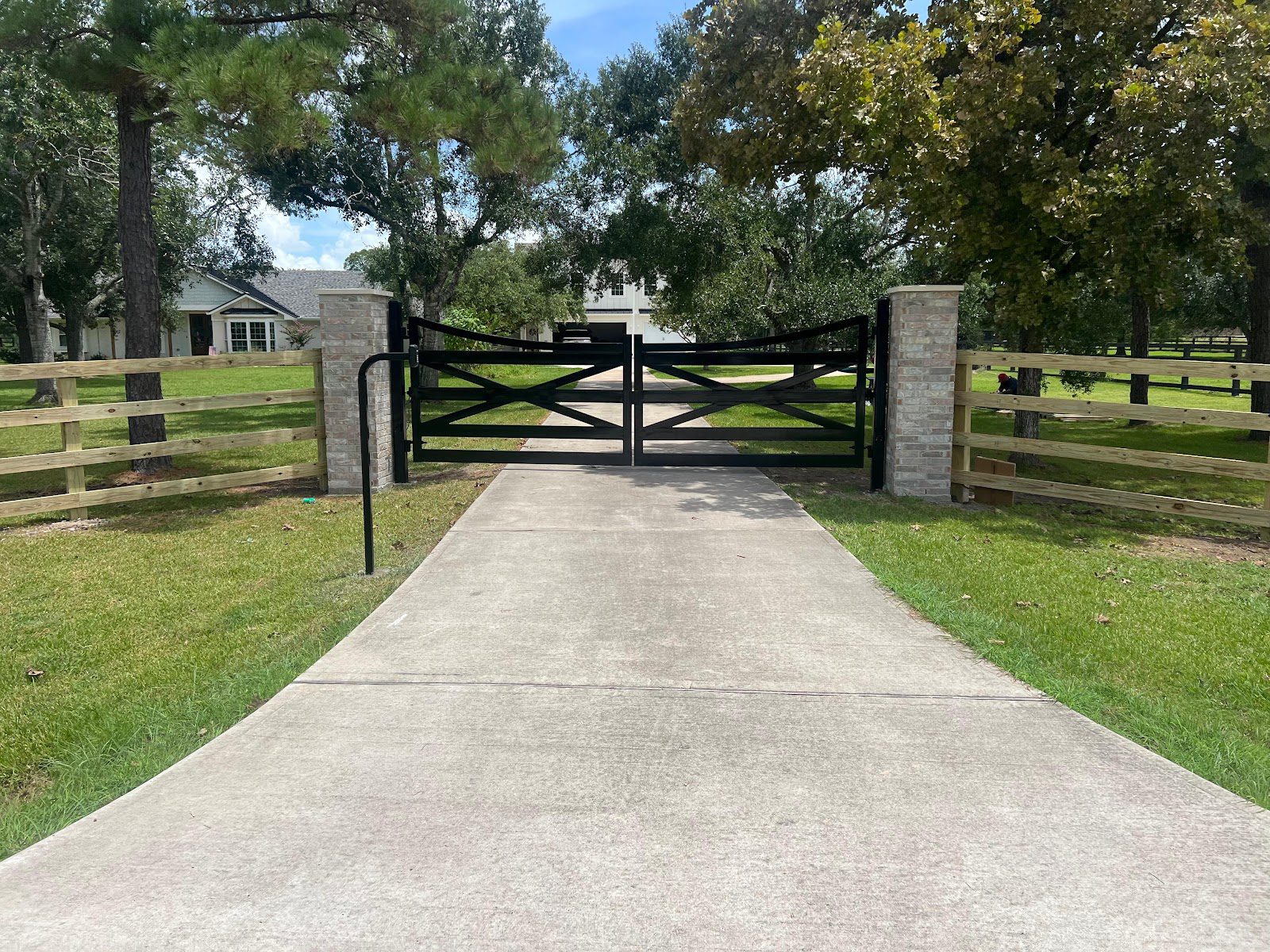 A custom black steel double swing gate sits closed between brick columns, its two X-braced leaves meeting symmetrically at the center of the drive.