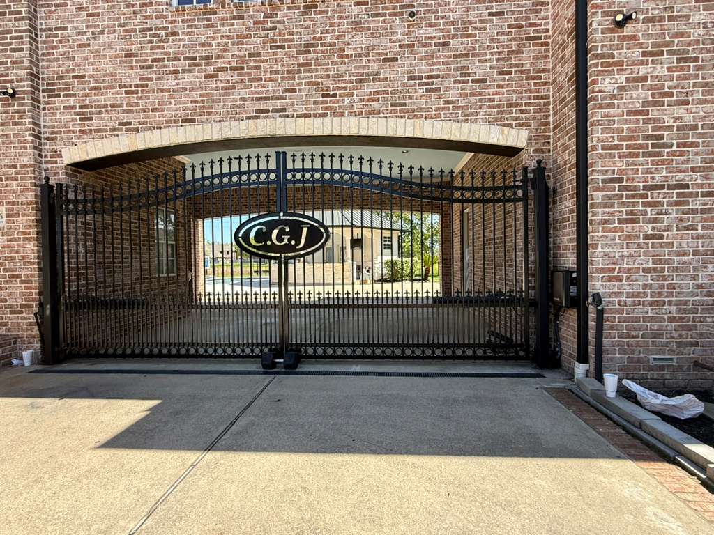 An ornate black wrought-iron double-swing gate driven by an automatic operator opens beneath a finished brick porte-cochere entrance.