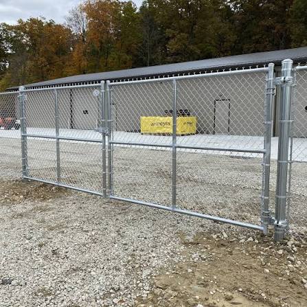 Galvanized silver chain-link with a diagonally braced gate section closes off a graveled work area beside a metal-clad commercial building, the same durable, economical enclosure detail we use to screen dumpsters and HVAC units behind Houston-area retail centers.