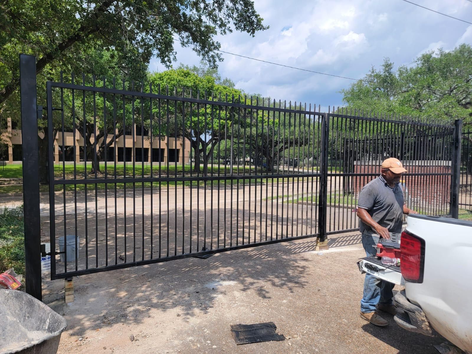 A Mustang Fencing technician works at a wide black steel spear-top double swing gate on install day at a shaded commercial entrance.