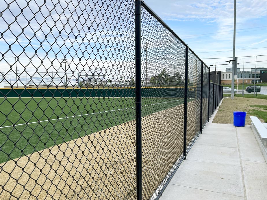 A black vinyl-coated chain-link perimeter fence on a concrete curb running the length of a turf ball field with a padded outfield wall.