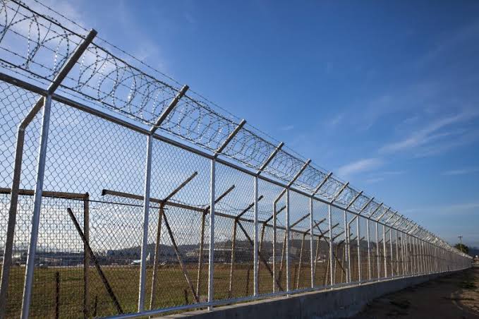 A hardened galvanized chain-link perimeter topped with concertina coils and angled barbed-wire outriggers locks down an industrial site under clear skies.