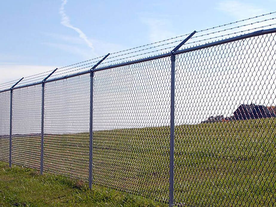 A tall galvanized chain-link fence topped with three strands of angled barbed wire runs along a grassy slope. It's the rugged, cost-effective perimeter Mustang installs around contractor lots and equipment yards near US-59 in Richmond.