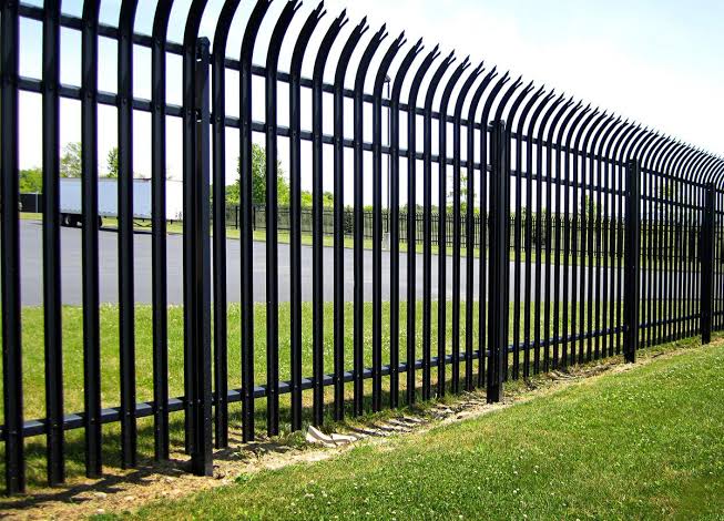 Black ornamental steel fencing with curved anti-climb picket tops encloses a commercial lot with a trailer parked behind it. That polished perimeter dresses up a retail center or office-park parking area in Tomball while keeping it secure.