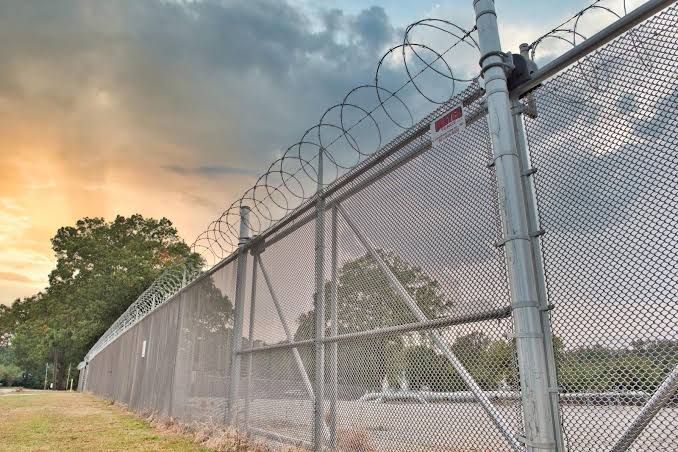 A galvanized security perimeter topped with concertina razor wire silhouetted against a sunset sky.