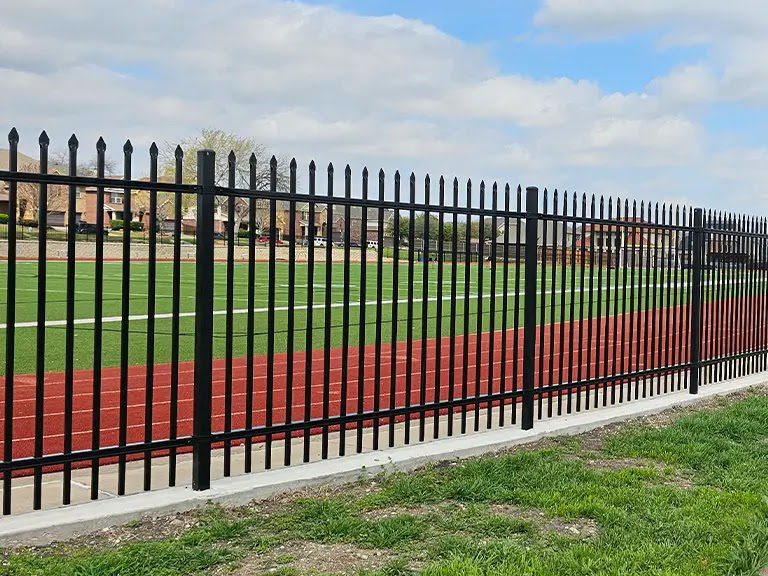 A black ornamental steel spear-top fence encloses a campus athletic complex, its open pickets keeping the grounds fully visible from the entrance drive.