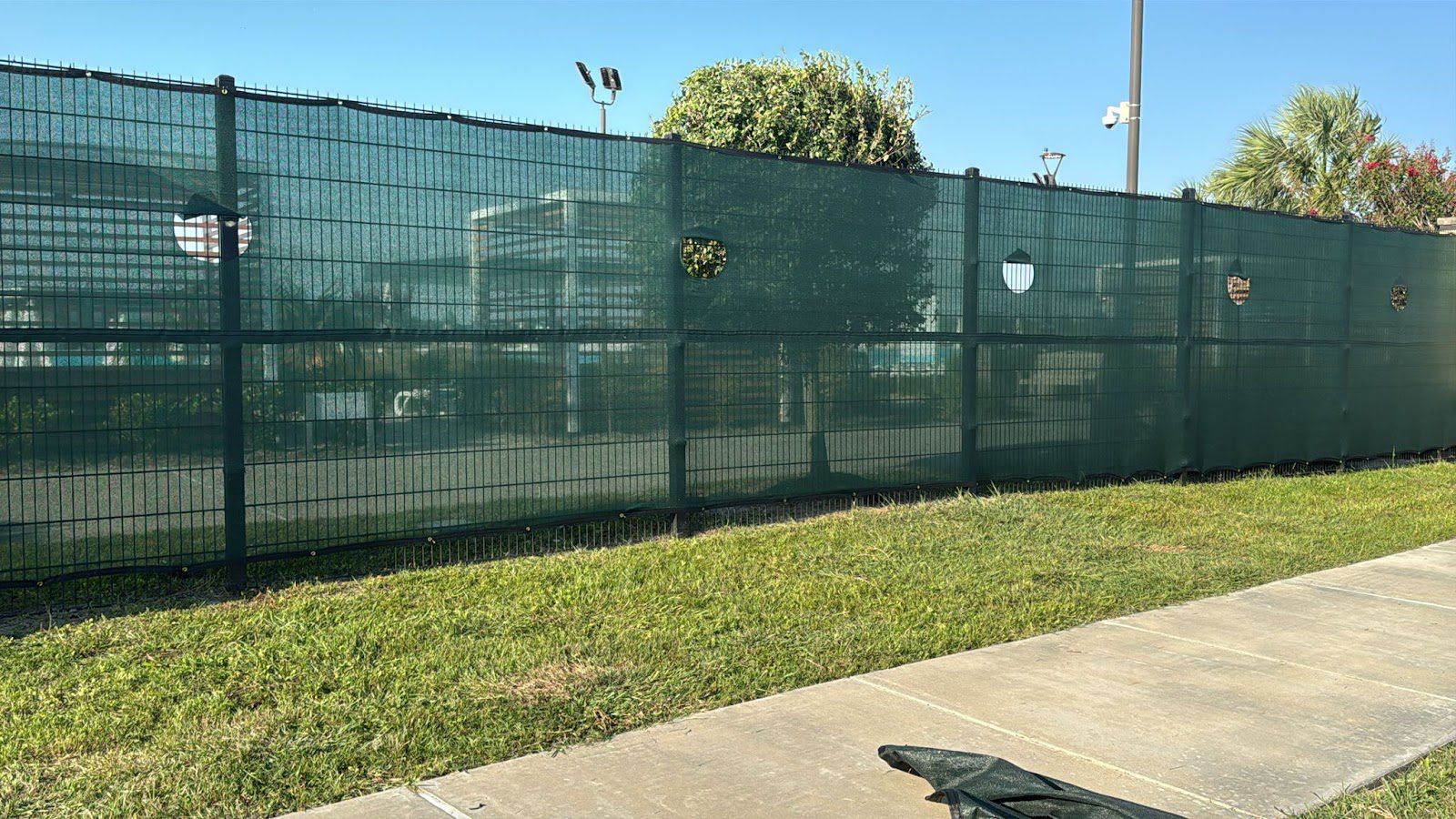 A welded steel mesh panel fence fitted with a green privacy wind screen shielding the sightline along a commercial parking area.