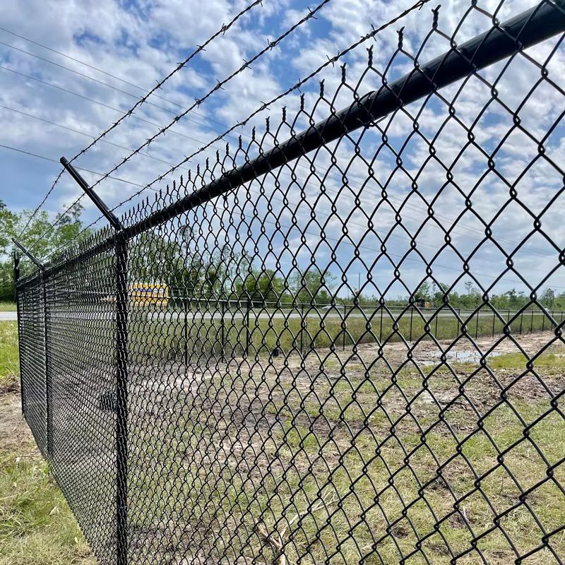 A vinyl-coated commercial chain-link perimeter fence topped with three-strand barbed wire securing an open industrial lot.