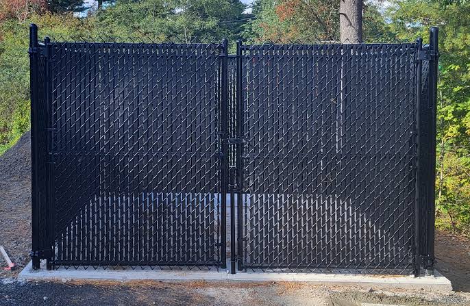 Black chain-link double gate with privacy slats woven through the mesh, closing off the gap at a commercial lot entrance.