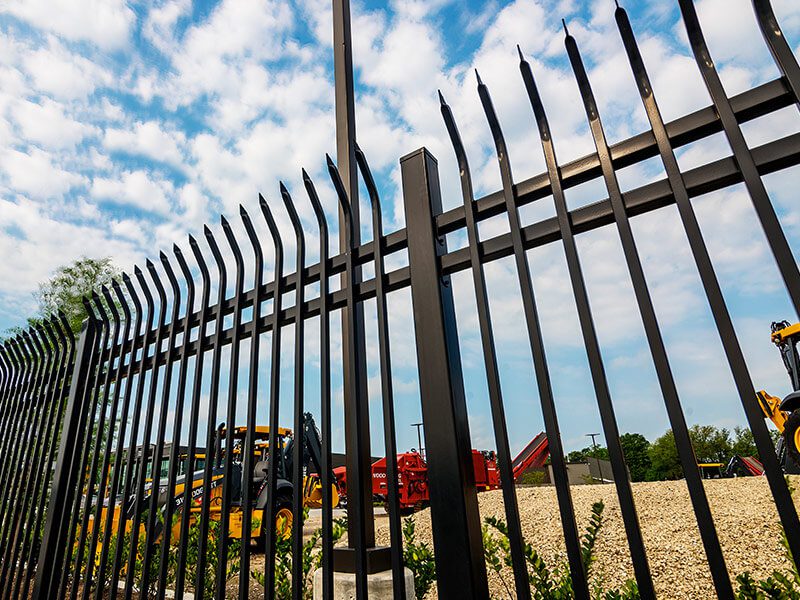 Skid-steers and heavy equipment stage just inside a tall black ornamental steel security fence, holding the working yard secure right at the perimeter.