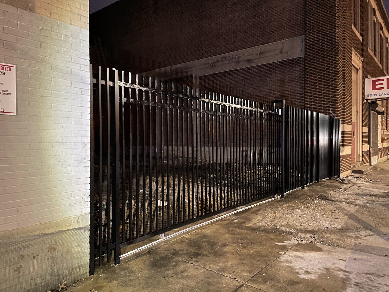 Completed black steel picket slide gate and matching fence beside a warehouse building, photographed at night